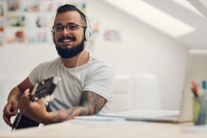 Man with acoustic guitar writing new song in his home recording studio. Using laptop and writing lyrics or music notes. Wearing headphones and playing guitar. Singing and smiling.